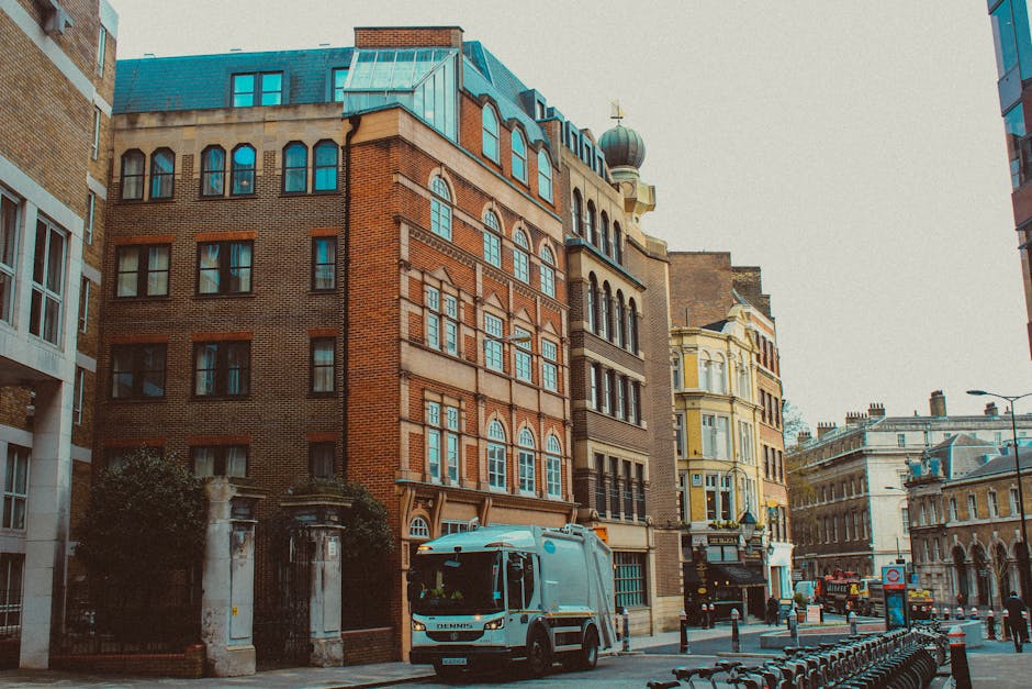 A city street scene featuring a row of historic multi-story buildings with a mix of brickwork and decorative architectural details, including arched windows and a small dome with a cross on top of one structure, indicating a religious or notable building. In the foreground, a white rubbish collection truck with blue and orange markings is parked along the curb, ready for waste collection. The street surface shows a narrow pavement lined with metal bike racks, and in the background, additional buildings and street furniture, such as lampposts and traffic signs, are visible under an overcast sky. The environment suggests an urban area where private waste collection services, like those from rubbishcollectionfinsburypark.co.uk, could facilitate on-site rubbish removal for local residents or businesses, especially in contrast to traditional municipal collection methods. The scene remains neutral and professional, with no human activity visible, emphasizing the operational context of rubbish handling and urban waste management.