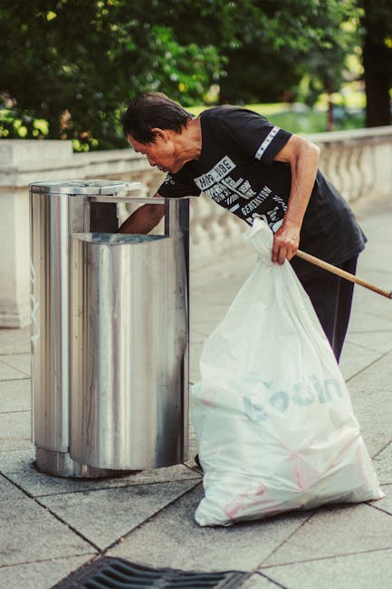 A middle-aged man with dark hair wearing a black t-shirt with white graphics is shown disposing of waste into a modern, silver, stainless steel public bin on a paved sidewalk. The bin has a rounded, cylindrical shape with a vertical opening on the front, designed for easy access to dispose of waste. The man holds a large, white plastic bag filled with rubbish in his left hand, while his right arm is extended directly into the bin, indicating he is actively depositing waste. The background features lush green trees and a stone balustrade, suggesting a park or outdoor public space. The natural daylight illuminates the scene evenly, capturing the textures of the man's clothing, the metallic finish of the bin, and the paved surface beneath. This scene illustrates community efforts towards proper rubbish disposal, representative of private or alternative waste handling services such as those offered by rubbish collection companies like rubbishcollectionfinsburypark.co.uk, emphasizing the importance of maintaining clean urban outdoor environments.