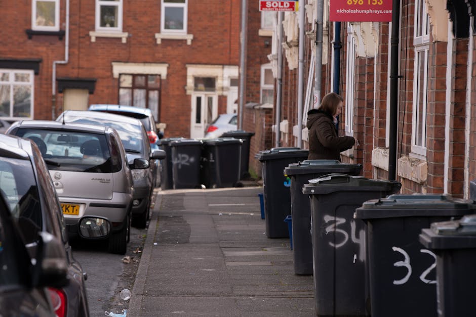 A row of traditional Victorian terraced houses with bay windows, brick facades painted in neutral tones, and tiled roofs with chimney stacks is visible in the background. In the foreground, there are two black public recycling bins placed on a grassy verge beside a paved footpath, with a large tree with fresh, bright green leaves extending its branches over the scene from the left side. The sky above is partly cloudy with patches of blue peeking through, creating a natural daylight atmosphere. The scene is set in a residential area with neatly maintained gardens and a low wooden fence separating the houses from the grassy area with the bins, representing a typical suburban environment that might be associated with alternative waste handling or private rubbish collection services in an urban setting. The overall setting suggests a peaceful neighbourhood with well-maintained infrastructure and clear pedestrian pathways.