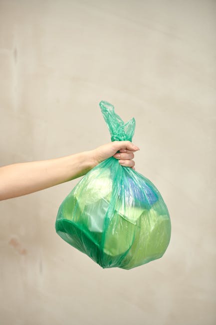 A person’s arm is visible extending from the left side of the image, holding a green plastic rubbish bag by its twisted top. The bag is filled with refuse, appearing slightly bulging and tied off at the top, with some translucent, crinkled plastic extending upward. The background features a plain, neutral-colored wall with subtle variations in shading, suggesting an indoor or outdoor setting with soft, diffuse lighting. The scene emphasizes the act of informal waste disposal, which may relate to private rubbish handling or alternative collection methods outside standard municipal services, reflecting typical rubbish removal activities supported by companies such as rubbishcollectionfinsburypark.co.uk.