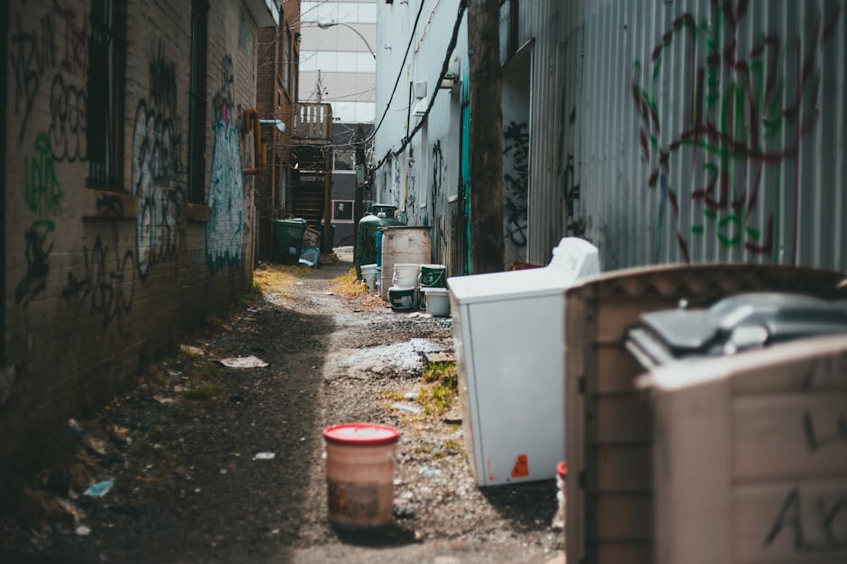 A narrow alleyway between two buildings lined with graffiti-covered brick and corrugated metal walls. The ground is uneven with patches of dirt, mud, and scattered debris. Several large green plastic bins, a white washing machine, and a brown trash container are situated along the right side of the alley, indicating private waste disposal. Further down, more green bins and a small plastic container are visible, with a staircase leading up to an upper floor at the end of the alley. The lighting appears natural, with daylight filtering in from the open end, highlighting the textures of the graffiti, rusted metal surfaces, and muddy ground. The scene suggests an environment where independent rubbish collection or on-site clearance is necessary, with visible signs of informal waste management practices typical of alleyway refuse accumulation. Such settings often require professional rubbish removal services to manage waste effectively in tight, urban spaces.