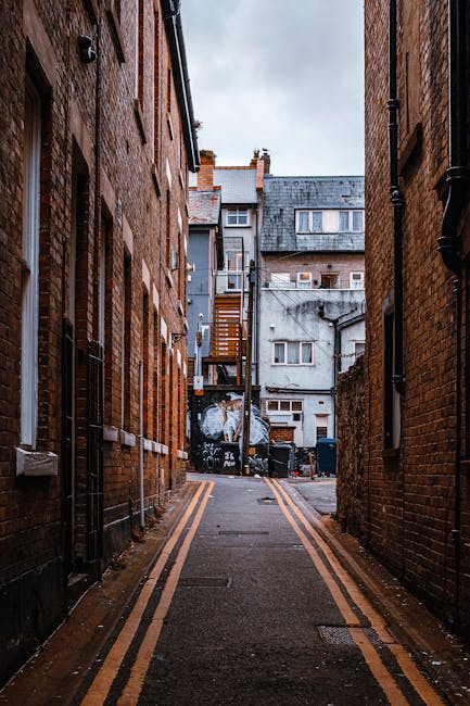 A narrow alleyway situated between two brick buildings in an urban setting, with weathered red brick walls on either side and a dark asphalt surface marked with double yellow lines running along the edges. In the distance, there is a small courtyard area with a grey, multi-storey building featuring a mix of concrete and brick finishes, small rectangular windows, and external staircases. Several black and grey waste bins are lined up along the back wall of the courtyard, and there are patches of graffiti on the lower part of the wall. The scene is illuminated by natural daylight under an overcast sky, creating a muted, neutral tone. The alley evokes a setting suitable for private waste collection or on-site clearance, in line with independent rubbish removal services. The surroundings are typical of urban alleyways used for waste disposal, highlighting the context of alternative rubbish handling outside of municipal waste systems.