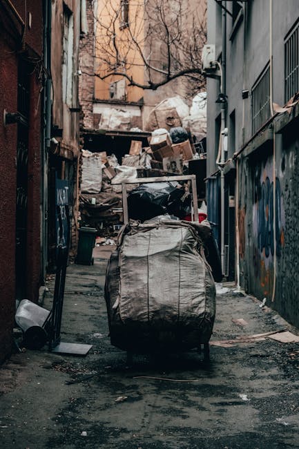A narrow alleyway between two buildings lined with graffiti-covered brick and corrugated metal walls. The ground is uneven with patches of dirt, mud, and scattered debris. Several large green plastic bins, a white washing machine, and a brown trash container are situated along the right side of the alley, indicating private waste disposal. Further down, more green bins and a small plastic container are visible, with a staircase leading up to an upper floor at the end of the alley. The lighting appears natural, with daylight filtering in from the open end, highlighting the textures of the graffiti, rusted metal surfaces, and muddy ground. The scene suggests an environment where independent rubbish collection or on-site clearance is necessary, with visible signs of informal waste management practices typical of alleyway refuse accumulation. Such settings often require professional rubbish removal services to manage waste effectively in tight, urban spaces.