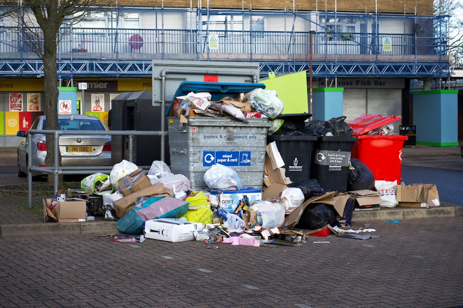 In an outdoor urban setting, a large pile of mixed waste materials is gathered around multiple overflowing rubbish bins on a paved surface. The waste includes crumpled cardboard boxes, paper packaging, plastic bags, and other household trash which spill onto the ground. Several black and red plastic rubbish bags sit on and around the bins, with some torn open, revealing its contents. The bins are labeled for mixed paper, cardboard, and general waste, indicating private or independent waste handling that may complement local rubbish collection services. Behind the waste, there is a vehicle parked nearby, partially obscured by a metal railing, with a building structure in the background featuring parking spaces and a storefront with signage. The environment appears to be a roadside or parking area, with a leafless tree on the left and a clear sky above, suggesting daytime. This scene illustrates the need for effective rubbish removal and waste management solutions, especially during on-site clearance or private disposal activities, as represented on the Finsbury Park rubbish collection webpage.