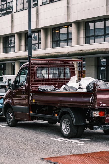 A dark red mini truck with an open bed loaded with various bulky waste items, including plastic bags, cardboard boxes, and other miscellaneous refuse, parked on a city street in front of a modern office building with large windows and concrete exterior. The truck features a metal mesh partition behind the cab, and its tires are visible beneath the bed. The surrounding environment includes a black pole and a white vehicle partially visible nearby, suggesting a typical urban setting for waste collection or private rubbish removal. The scene reflects an instance of on-site clearance or alternative waste handling, potentially managed by a private rubbish disposal service like rubbishcollectionfinsburypark.co.uk. The lighting appears natural, with diffuse daylight illuminating the scene, emphasizing the textures and colours of the waste materials and the vehicle.