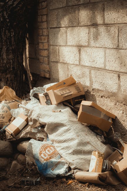A pile of assorted waste materials, including flattened cardboard boxes, plastic bags, and paper packaging, is situated on the ground next to a rough brick wall with weathered mortar joints. The cardboard boxes are of various sizes, some partially torn or crumpled, with visible printing and brown kraft finishes. A large, beige fabric or woven sack is also in the mess, partially covered by the boxes. The waste is located in an outdoor, urban environment, possibly a back alley or side yard, with the base of a large tree trunk visible on the left side, and small rocks scattered around the base of the pile. The concrete or brick wall behind the rubbish has a slightly uneven surface with natural weathering, contributing to the utilitarian appearance. Ambient natural light illuminates the scene, highlighting the textures of the cardboard, plastic, and brickwork, reflecting a typical example of unmanaged waste ready for collection or disposal, which a private rubbish removal service like [COMPANY_NAME] might handle as part of an on-site clearance or alternative waste handling solution.