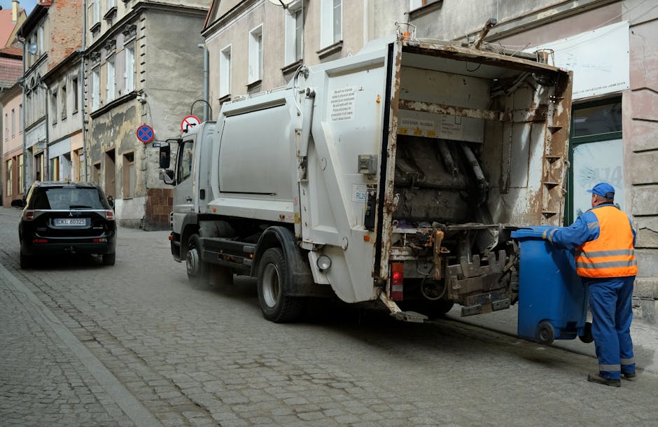 A dark red mini truck with an open bed loaded with various bulky waste items, including plastic bags, cardboard boxes, and other miscellaneous refuse, parked on a city street in front of a modern office building with large windows and concrete exterior. The truck features a metal mesh partition behind the cab, and its tires are visible beneath the bed. The surrounding environment includes a black pole and a white vehicle partially visible nearby, suggesting a typical urban setting for waste collection or private rubbish removal. The scene reflects an instance of on-site clearance or alternative waste handling, potentially managed by a private rubbish disposal service like rubbishcollectionfinsburypark.co.uk. The lighting appears natural, with diffuse daylight illuminating the scene, emphasizing the textures and colours of the waste materials and the vehicle.
